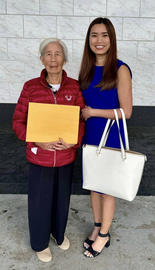A professional young woman in a blue dress, identified as a licensed immigration assistance provider from Viet US Guide, stands proudly next to a new U.S. citizen, an elderly lady holding a yellow envelope, contains her naturalization documents. They are both smiling and standing in front of a marble wall, celebrating the older woman's successful journey to American citizenship.