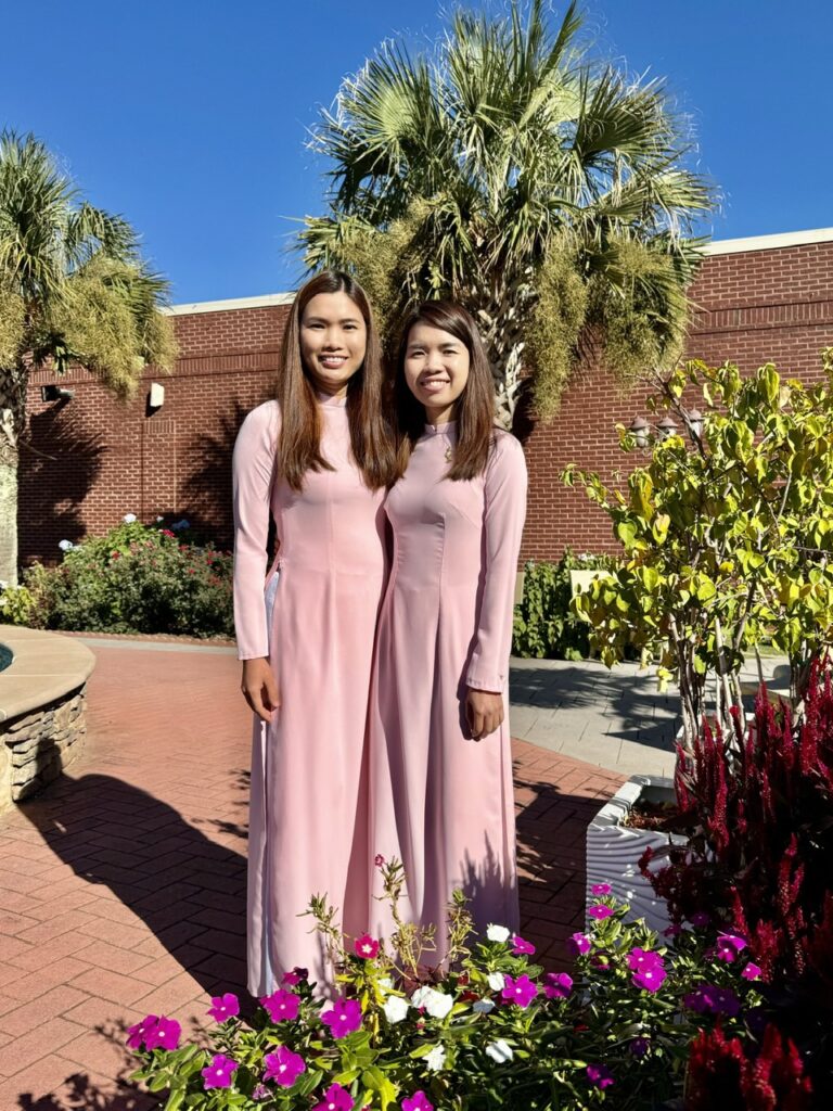 Two smiling women in pink áo dài standing before palm trees and a flower garden at a Vietnamese cultural event in Georgia.