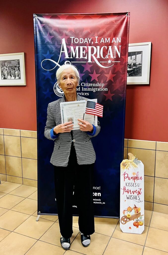 Newly naturalized elderly Vietnamese woman proudly displays her U.S. naturalization certificate and flag in front of a "Today, I am an American" banner.