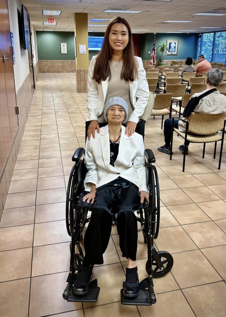 An elderly Vietnamese woman in a wheelchair, proudly naturalized as a U.S. citizen with the support of Viet US Guide, with her family member standing beside her in celebration.